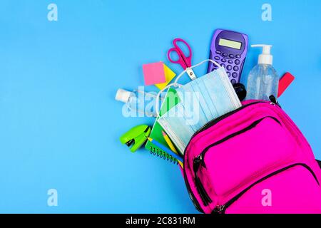 Sac à dos plein de fournitures scolaires et articles de prévention COVID 19. Vue de dessus, en renversant sur un fond bleu. Retour à l'école pendant la pandémie concept. Banque D'Images