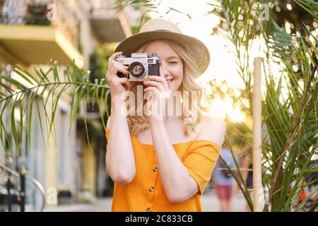 Jolie fille blonde souriante dans un chapeau prendre des photos sur un appareil photo rétro dans la rue Banque D'Images