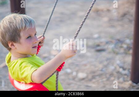 Joyeux petit garçon blond est sur une balançoire, jouant dehors sur le terrain de jeu dans le parc. Garderie, activité, joies simples de l'enfance Banque D'Images