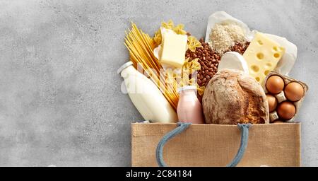 Sac de shopping rempli de produits laitiers frais et de glucides tels que fromage, pâtes, baguette, haricots, œufs et lait sur fond gris texturé Banque D'Images