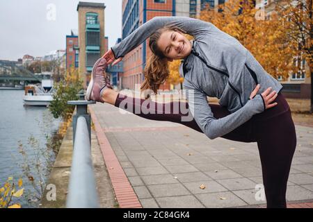 Jolie fille sportive souriante s'étirant tranquillement pendant l'entraînement dans la rue de la ville Banque D'Images