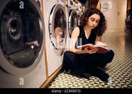 Belle fille brunette lire attentivement le livre en attente de lavage dans la blanchisserie moderne en libre-service Banque D'Images