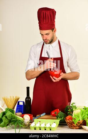 Chef avec visage occupé tient un bol et fouetter sur fond blanc. Ustensiles de cuisine et concept de cuisine. Cuisiner travaille dans la cuisine près de la table avec des légumes et des outils. Homme dans le chapeau de cuisinier et le tablier tient les ustensiles de cuisine Banque D'Images