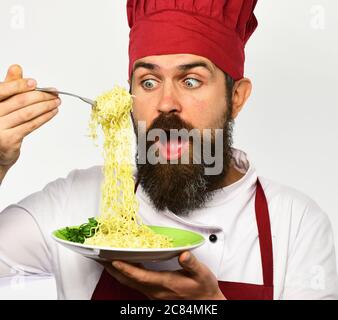 Cuire avec le visage choqué dans l'uniforme bordeaux tient la fourchette et l'assiette. Concept de cuisine de restaurant. Le chef mange des nouilles italiennes ou asiatiques. L'homme avec la barbe tient un plat savoureux sur fond blanc. Banque D'Images