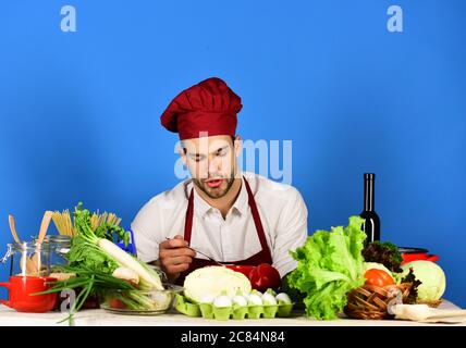 Ustensiles de cuisine et concept de cuisine. L'homme en cuisine, le chapeau et le tablier, mange de la soupe à table. Cuisiner travaille dans la cuisine près de la table avec des légumes et des outils. Chef avec visage occupé tient la cuillère sur fond bleu. Banque D'Images