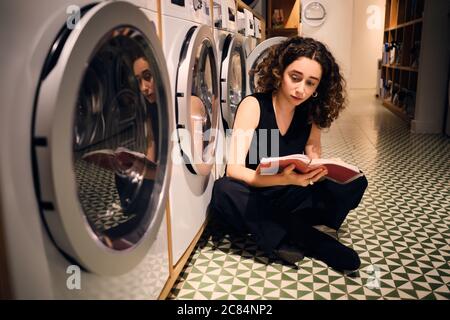 Jolie fille de brunette lecture réfléchie livre attente lavage dans la blanchisserie moderne self-service Banque D'Images