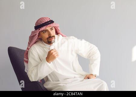 Un homme d'affaires arabe pensif assis sur une chaise pense qu'il regarde sur un fond gris. Portrait d'un homme arabe attrayant. Banque D'Images