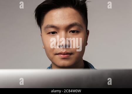 Gros plan sur un beau homme asiatique concentré travaillant avec un ordinateur portable isolé sur fond gris Banque D'Images