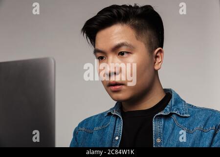 Gros plan sur un beau homme asiatique concentré travaillant avec un ordinateur portable isolé sur fond gris Banque D'Images