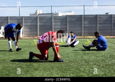 Multiethnique masculin cinq joueurs de football de côté portant un entraînement de bande d'équipe sur un terrain de sport au soleil, en se réchauffant de leurs lacets. Banque D'Images