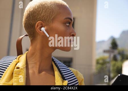 Femme alternative de course mixte avec des cheveux blond courts dehors et autour de la ville pendant une journée ensoleillée, portant des écouteurs sans fil et un sac à dos, en regardant loin. Banque D'Images