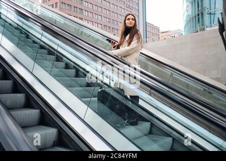 Jeune femme d'affaires décontractée avec valise sur escalier roulant extérieur Banque D'Images