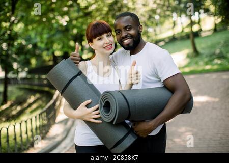 Jeune couple multiethnique souriant, garçon africain et fille caucasienne, portant des vêtements de sport, tenant des tapis de yoga, est prêt pour l'exercice dans le parc de la ville Banque D'Images