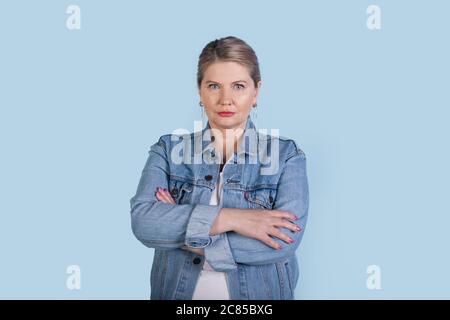 Femme caucasienne âgée sérieuse avec cheveux blonds portant un Jean manteau posant avec des mains croisées sur un mur bleu de studio Banque D'Images