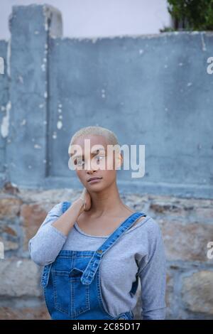 Femme alternative de course mixte avec cheveux blond courts portant des salopettes en denim dehors et autour de la ville par temps ensoleillé, debout près d'un mur et regardant un Banque D'Images