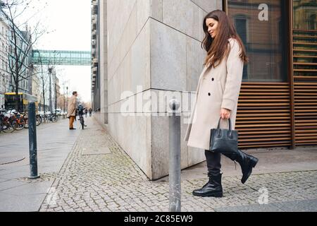 Vue latérale de l'élégante fille de brunette en manteau de rêve marchant dans la rue de la ville Banque D'Images