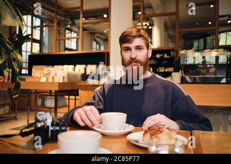 Jeune homme barbu attrayant regardant attentivement dans l'appareil photo ayant une pause café dans le café Banque D'Images