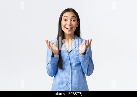 Bonne fille asiatique en pyjama bleu levant les mains et applaudir dans le plaisir, souriant émerveillé, écoutant de merveilleuses nouvelles, louant le bon travail Banque D'Images