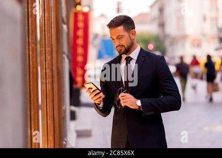 Un beau jeune homme d'affaires qui marche dans la rue et vérifie une vitrine tout en utilisant son smartphone. Banque D'Images