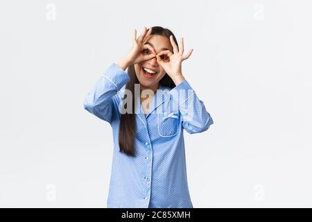 Bonne fille asiatique en pyjama bleu montrant de fausses lunettes gestuelle et souriant optimiste, s'amuser avec les frères et sœurs à la fête de la nuit, portant des jambies Banque D'Images