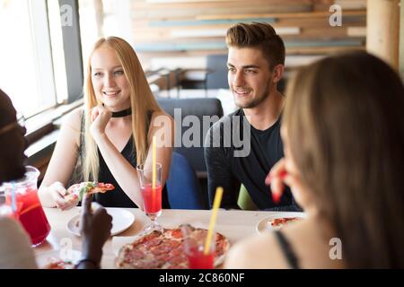 Groupe de jeunes gens de race mixte divers visitant un café élégant et bien éclairé, manger de la nourriture de rue et malsaine, apprécier la pizza et le cocktail de fruits, en Banque D'Images