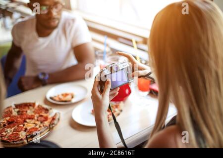Des amis multiraciaux heureux qui dégusterent une pizza au restaurant, tandis que l'un des plus fermes prend des photos de délicieux plats pour les télécharger sur les réseaux sociaux Banque D'Images