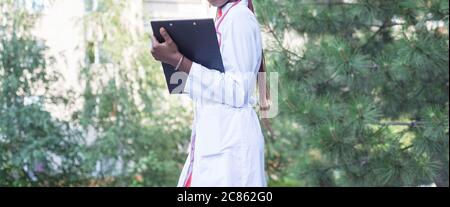 Une femme afro-américaine, âgée de vingt-sept ans, en manteau blanc, avec un phonendoscope, écrit un stylo dans un dossier pour les papiers. Sur un fond noir Banque D'Images