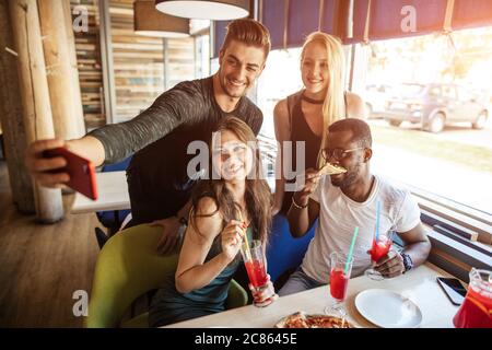Groupe multiethnique de jeunes étudiants joyeux s'amusant au café de la fête avec de savoureux fast food, des boissons, en prenant le selfie sur smartphone Banque D'Images