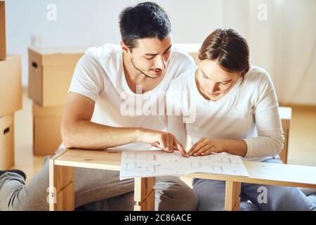 Le couple lit les instructions pour l'assemblage de meubles dans une nouvelle maison avec des boîtes mobiles sur l'arrière-plan Banque D'Images