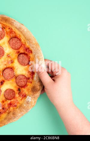 Image verticale avec la main d'une femme saisissant une pizza pepperoni. Vue de dessus avec une pizza poivrière fraîchement cuite sur fond vert menthe. Banque D'Images
