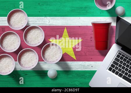 Drapeau du Suriname avec tasses de bière-pong en plastique et ordinateur portable sur table nationale en bois, vue sur le dessus. Jeu de pong de bière. Banque D'Images