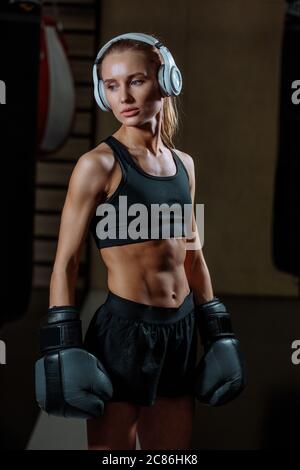 femme avec abs parfait portant des écouteurs et des vêtements noirs debout dans la salle de gym près du sac de poinçonnage Banque D'Images