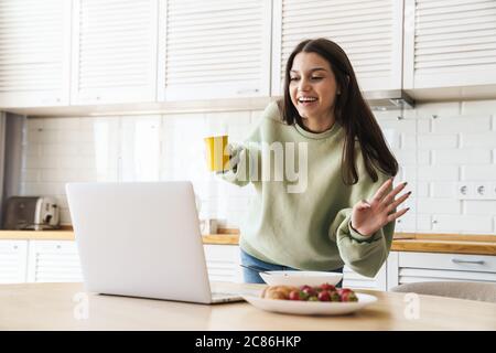 Photo de gaie belle femme agitant la main et utilisant l'ordinateur portable tout en prenant le petit déjeuner dans la cuisine moderne Banque D'Images