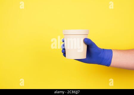 Main de l'homme dans des gants de protection jetables bleus sur fond jaune tenant un récipient à nourriture en papier rond - gobelet en carton pour soupe, crème glacée ou autre Banque D'Images