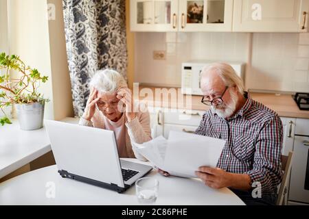 Couple d'âge mûr assis à la table de cuisine avec ordinateur portable regardant à travers les papiers financiers, ayant peu de confiture avec les cotisations de retraite. Banque D'Images