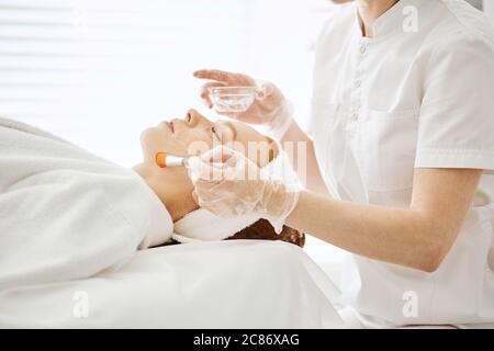 La cosmétologue féminine applique du gel hydrogélique sur une femme aux cheveux rouges, les yeux fermés étant couchés sur un lit médical. Jeune femme étant prête au traitement au laser. Banque D'Images