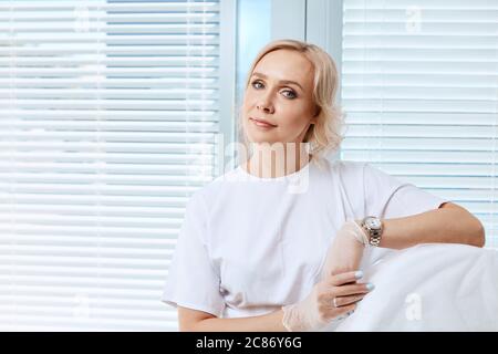 Portrait d'une femme blonde confiante en uniforme blanc souriant regardant l'appareil photo et posant dans un centre médical moderne assis contre le W Banque D'Images