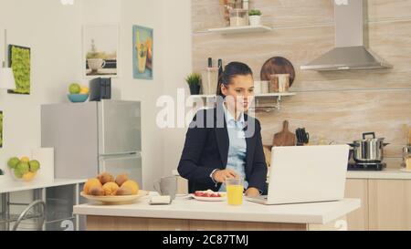 Jeune femme d'affaires dans la cuisine qui a un repas sain tout en parlant sur un appel vidéo avec ses collègues du bureau, en utilisant la technologie moderne et de travailler 24 heures sur 24 Banque D'Images