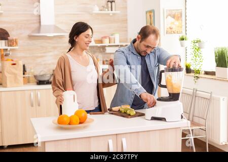 Un couple joyeux prépare un smoothie à l'aide d'un blender. Femme tenant le biberon dans la cuisine. Un style de vie sain et insouciant, une alimentation et la préparation du petit déjeuner dans un matin ensoleillé Banque D'Images