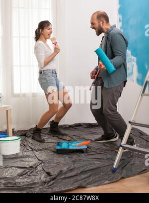 Fille chantant sur la brosse de peinture et de décoration d'appartement avec l'aide de mari gai. Rénovation d'appartements et construction de maisons tout en rénove et en améliorant. Réparation et décoration. Banque D'Images