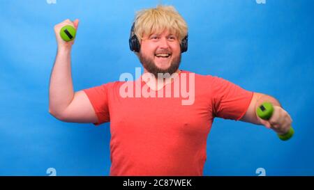 Drôle d'homme de graisse freaky dans une perruque avec des haltères faisant des exercices sur un fond bleu. Banque D'Images