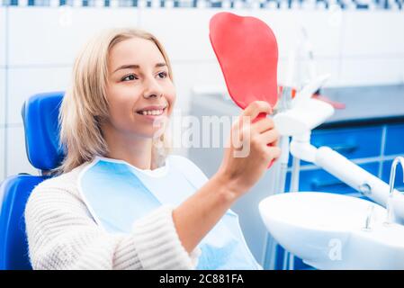 Fille blonde satisfaite regardant dans le miroir rouge après le travail du dentiste Banque D'Images