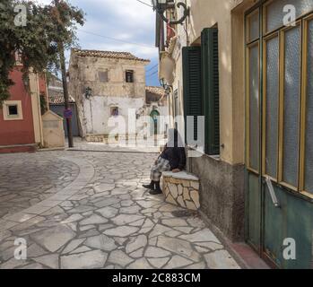 Grèce, corfou, Krini, 26 septembre 2018: Femme âgée en robe traditionnelle assise sur banc de pierre sur la place avec des maisons anciennes dans le village de Krini, tôt Banque D'Images