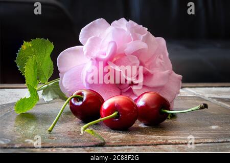 Gros plan de trois cerises rouges avec une rose rose et des feuilles vertes sur une surface brune. Photo macro, arrière-plan sombre, illustration Banque D'Images