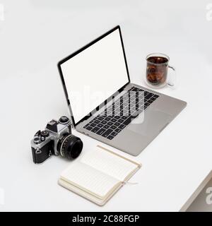 Photo rognée d'un ordinateur portable à écran vierge sur une table blanche avec carnet ouvert, appareil photo et tasse à café Banque D'Images