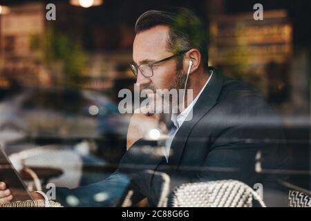 Homme d'affaires mature au café avec écouteurs regardant sa tablette numérique. Professionnel effectuant un appel vidéo depuis sa tablette tout en stit Banque D'Images