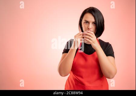 Femme employée au supermarché avec un tablier rouge et un t-shirt noir, boire du café de la tasse isolée sur fond rouge Banque D'Images