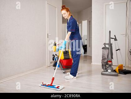 Belle jeune femme aux cheveux rouges regardant l'appareil photo à l'aide d'une vadrouille tout en nettoyant le sol dans la salle blanche. Concept de ménage Banque D'Images