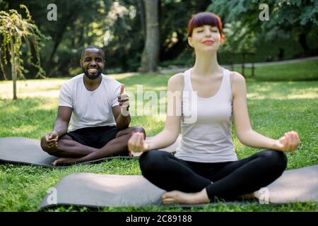 Jeune couple multiethnique se détendant en position lotus sur des tapis de coupe au parc. Caucasien jolie femme méditant avec les yeux fermés. Drôle Banque D'Images