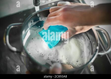 Un homme lave un pot dans l'évier de cuisine avec une éponge bleue et une mousse épaisse. Tâches ménagères. Ustensiles de cuisine. Banque D'Images